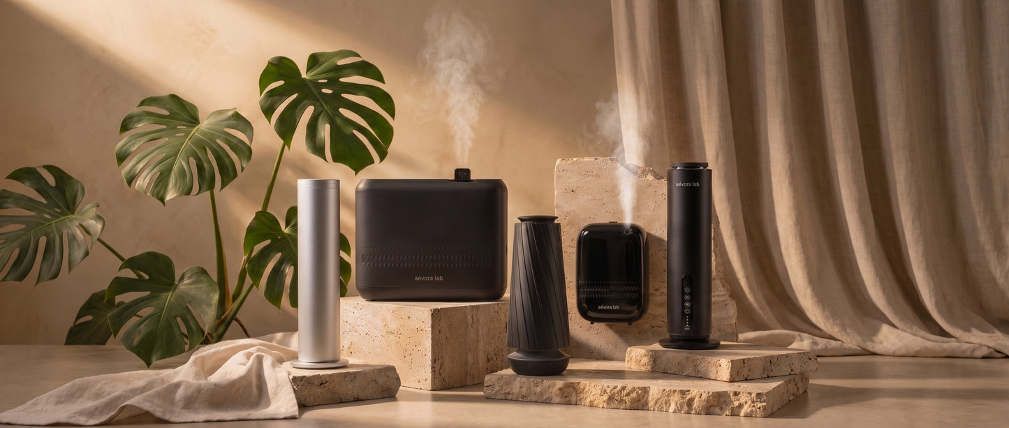 A group of modern home fragrance and wellness devices displayed together on stone pedestals in a warm, styled interior scene with draped beige curtains, a beige wall, and green monstera leaves. Visible products include a silver cylindrical diffuser, a black rectangular diffuser, a black cone-shaped diffuser, a black rectangular humidifier, and a black rectangular diffuser emitting mist.